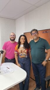 três pessoas em pé, uma mulher de cabelo cacheado e dois homens brancos